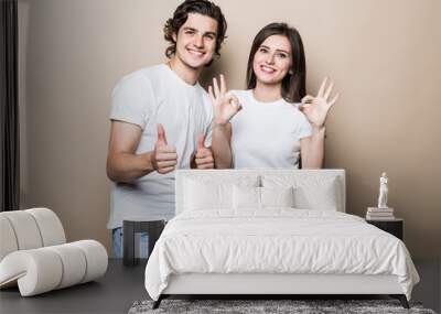 Portrait of beautiful people man and woman in basic clothing posing together at camera with thumbs up isolated over beige background Wall mural